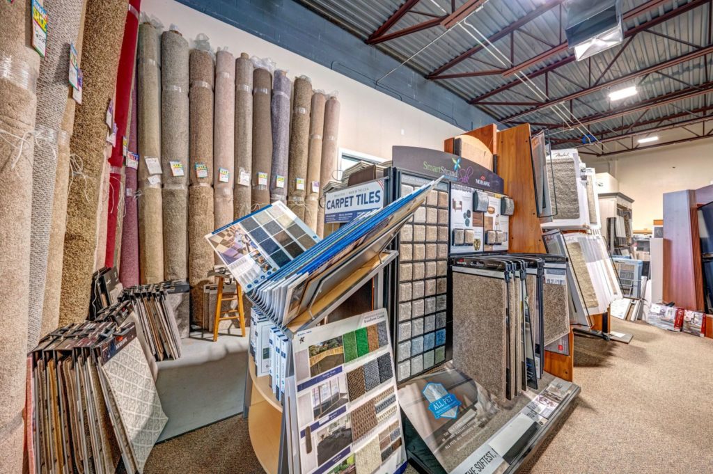 Showroom. In the front there is a carpet sample display and in the back there is large rolls of carpet.